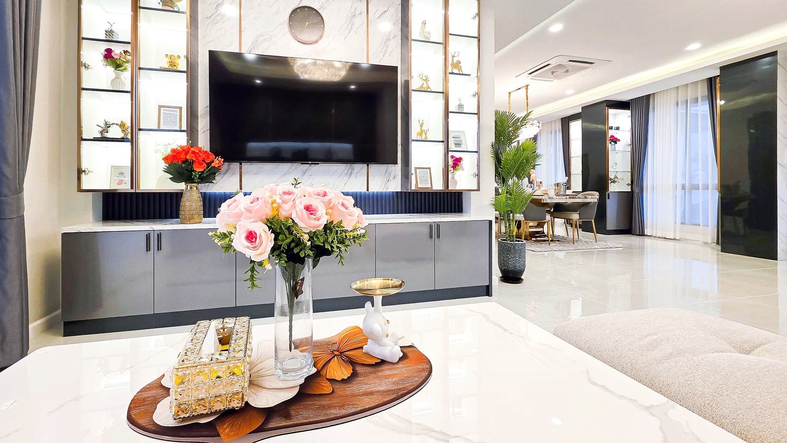 Bright modern living room with a large wall-mounted TV, marble accents, and glass display shelves filled with decor items.