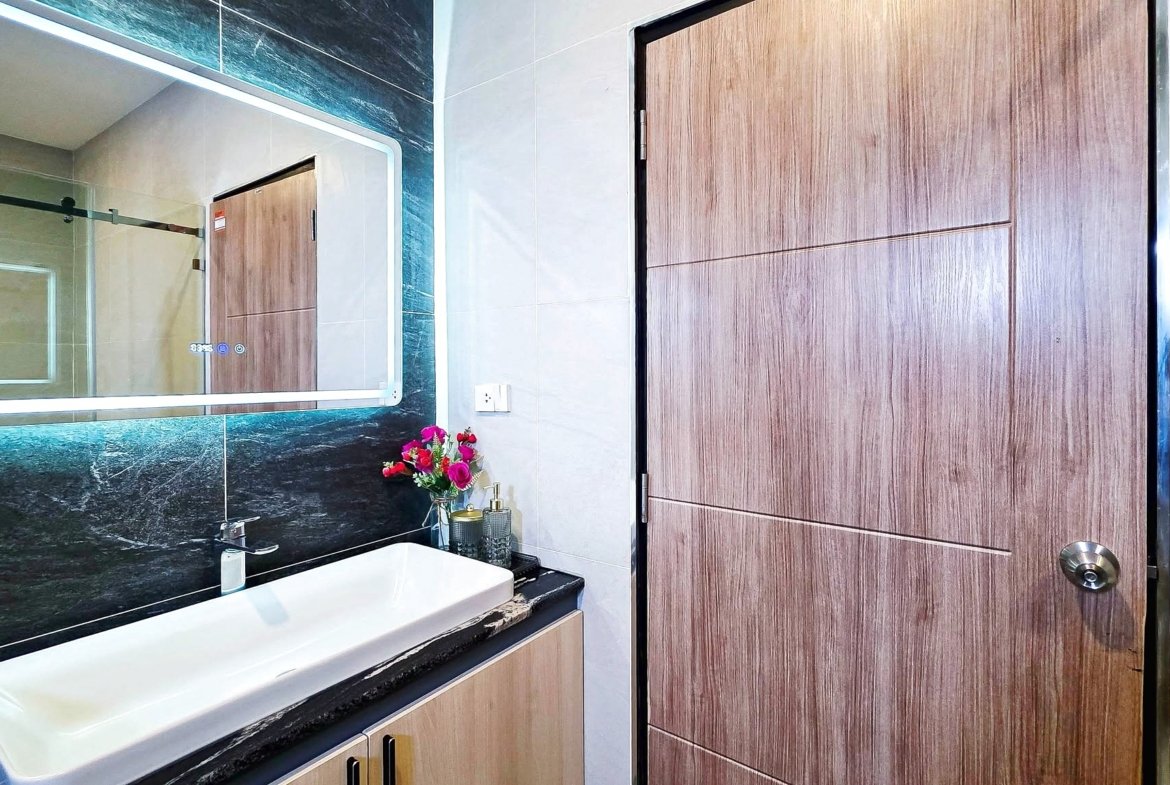 Modern bathroom with a white sink, black marble counter, illuminated backlit mirror, and a tall wood-paneled door; a small vase of flowers sits on the counter.