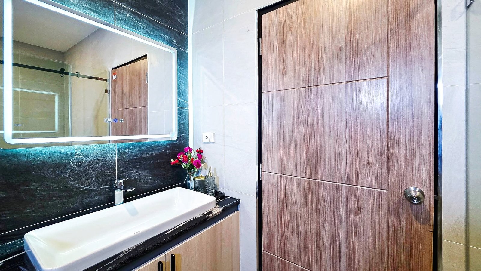 Modern bathroom with a white sink, black marble counter, illuminated backlit mirror, and a tall wood-paneled door; a small vase of flowers sits on the counter.