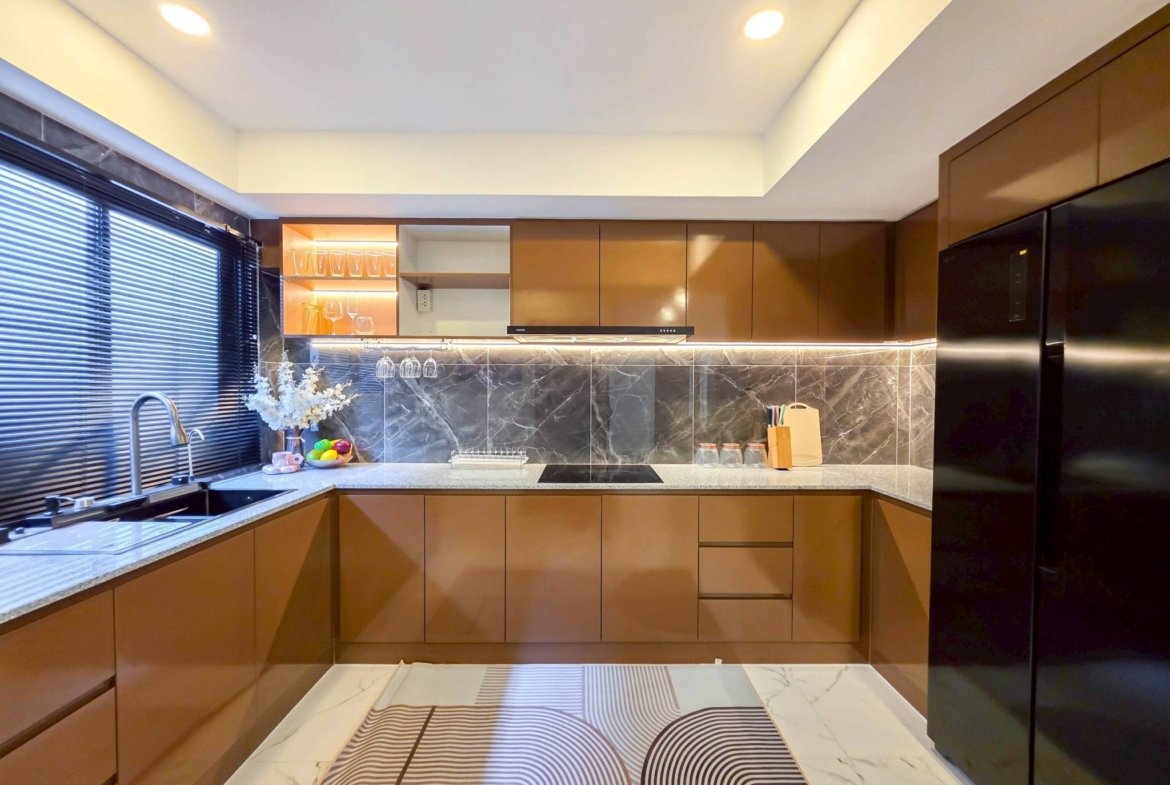 Modern L-shaped kitchen with glossy brown cabinets, marble backsplash, and a patterned marble floor under recessed ceiling lights. Window blinds along the left wall.
