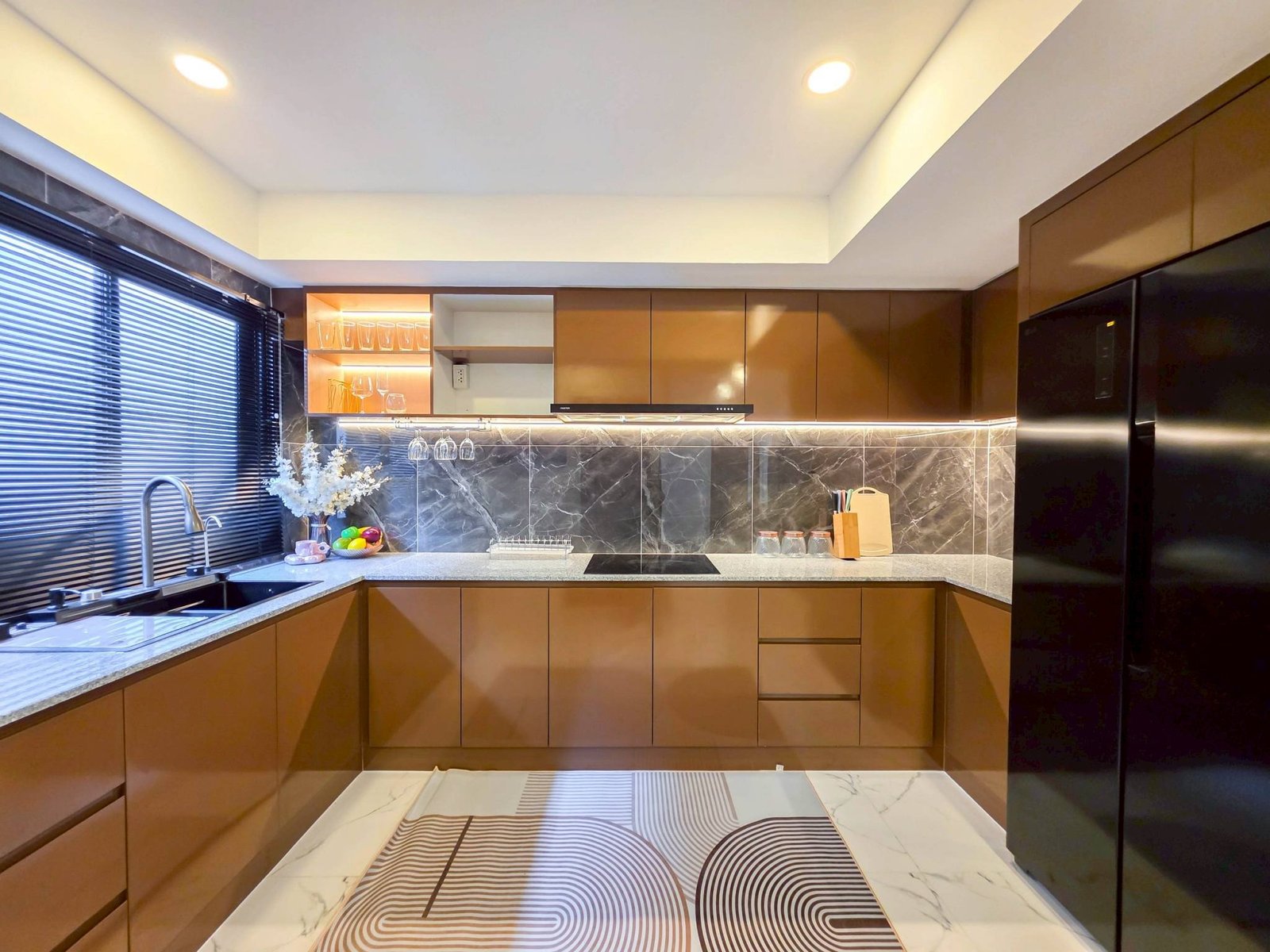 Pool_Villa_South_Pattaya_Sale_Dlh498_17 Modern L-shaped kitchen with glossy brown cabinets, marble backsplash, and a patterned marble floor under recessed ceiling lights. Window blinds along the left wall.