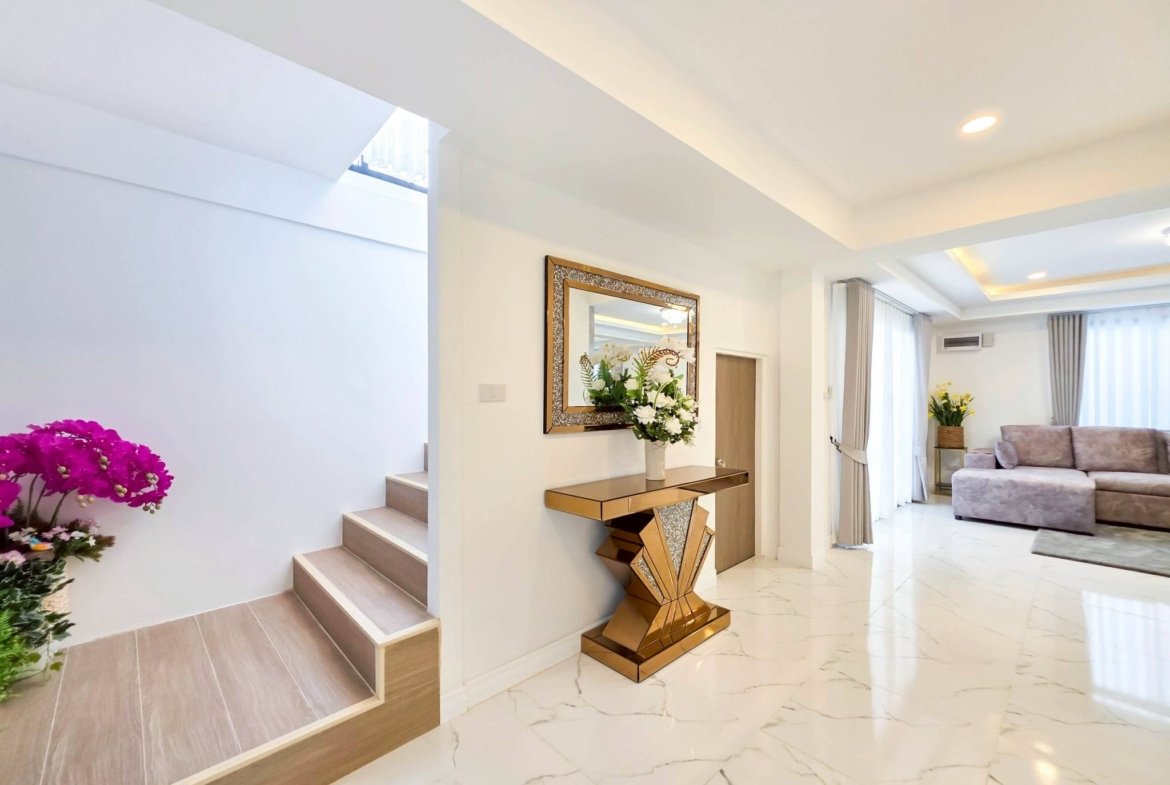 Modern living area with a wooden staircase on the left, a gold geometric console table, and a wall mirror, with pink flowers in the foreground and bright curtains in the background.