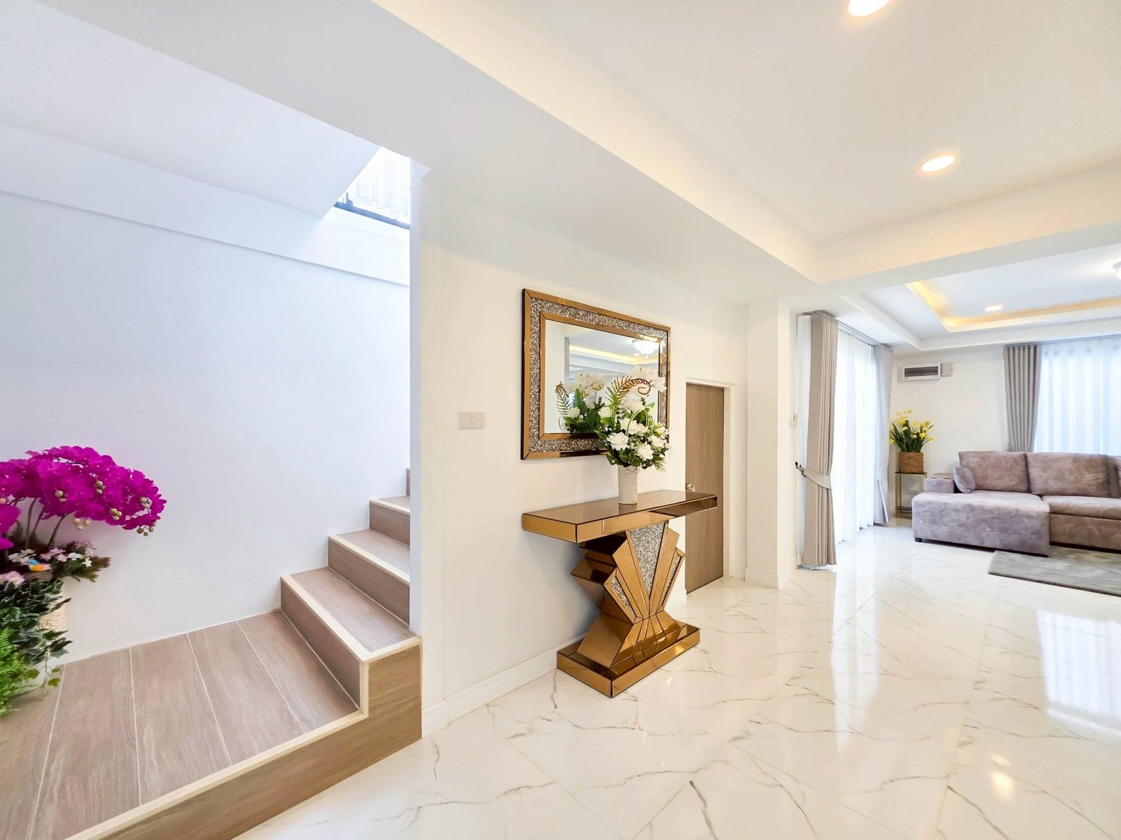 Pool_Villa_South_Pattaya_Sale_Dlh498_18 Modern living area with a wooden staircase on the left, a gold geometric console table, and a wall mirror, with pink flowers in the foreground and bright curtains in the background.