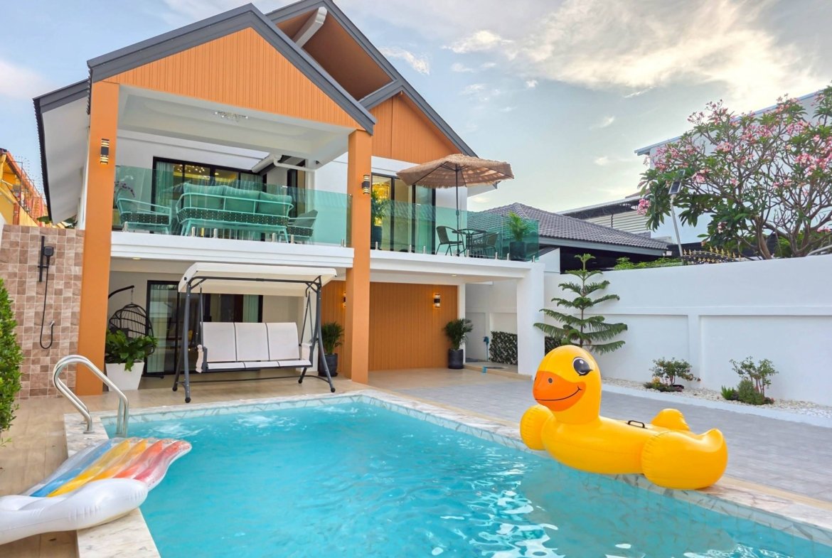 Modern two-story house with orange panels and a glass balcony overlooking a pool with a yellow inflatable duck in the water and patio furniture nearby.