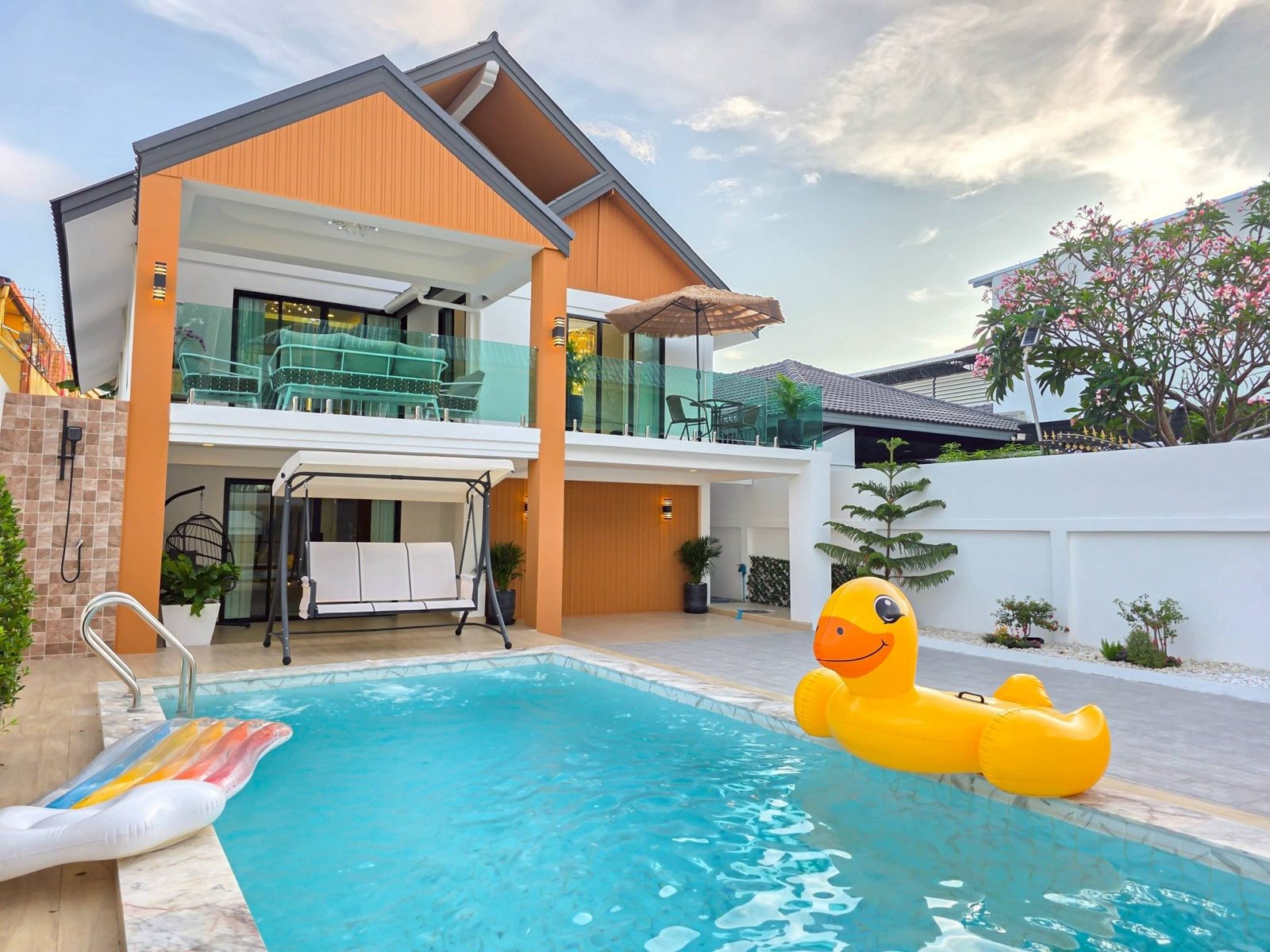 Pool_Villa_South_Pattaya_Sale_Dlh498_2 Modern two-story house with orange panels and a glass balcony overlooking a pool with a yellow inflatable duck in the water and patio furniture nearby.