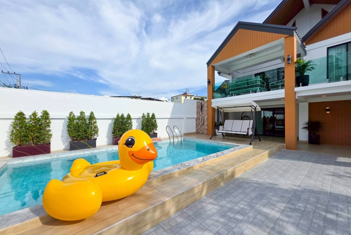 Residential pool deck with oversized yellow rubber duck floats in the water and modern outdoor furniture under blue sky, contemporary home exterior in the background.