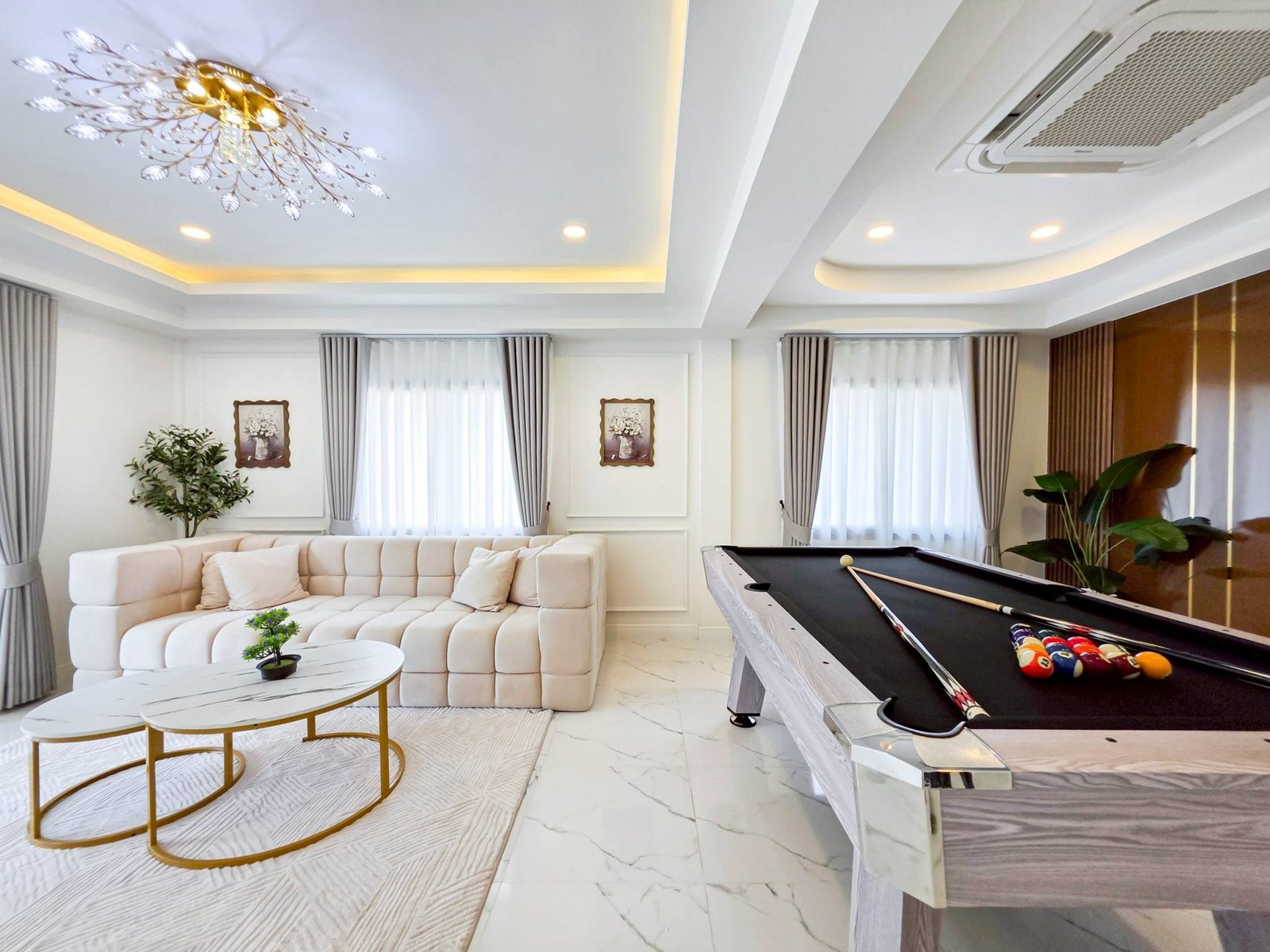Pool_Villa_South_Pattaya_Sale_Dlh498_4 Bright modern living room with a white tufted sectional sofa, glass-top coffee tables, and potted plants under a decorative chandelier.