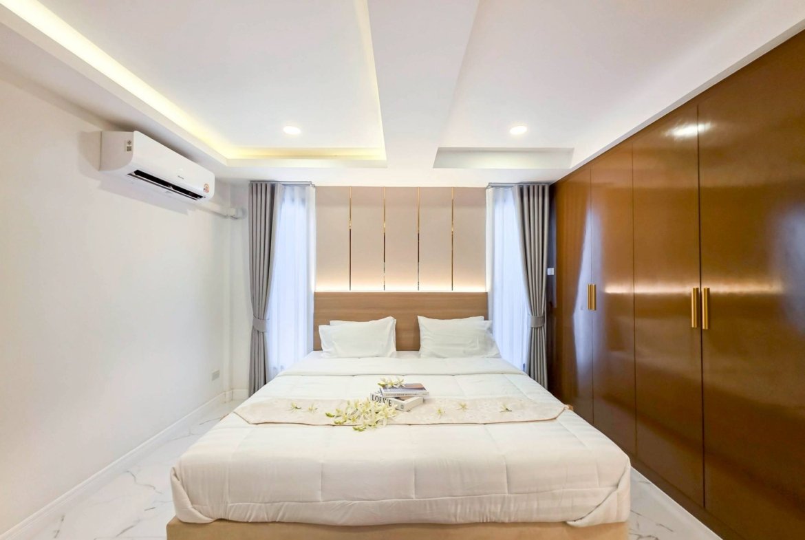 Hotel room with a large bed, white linens, and a flower-decorated runner; wooden wardrobe on the right and gray curtains.