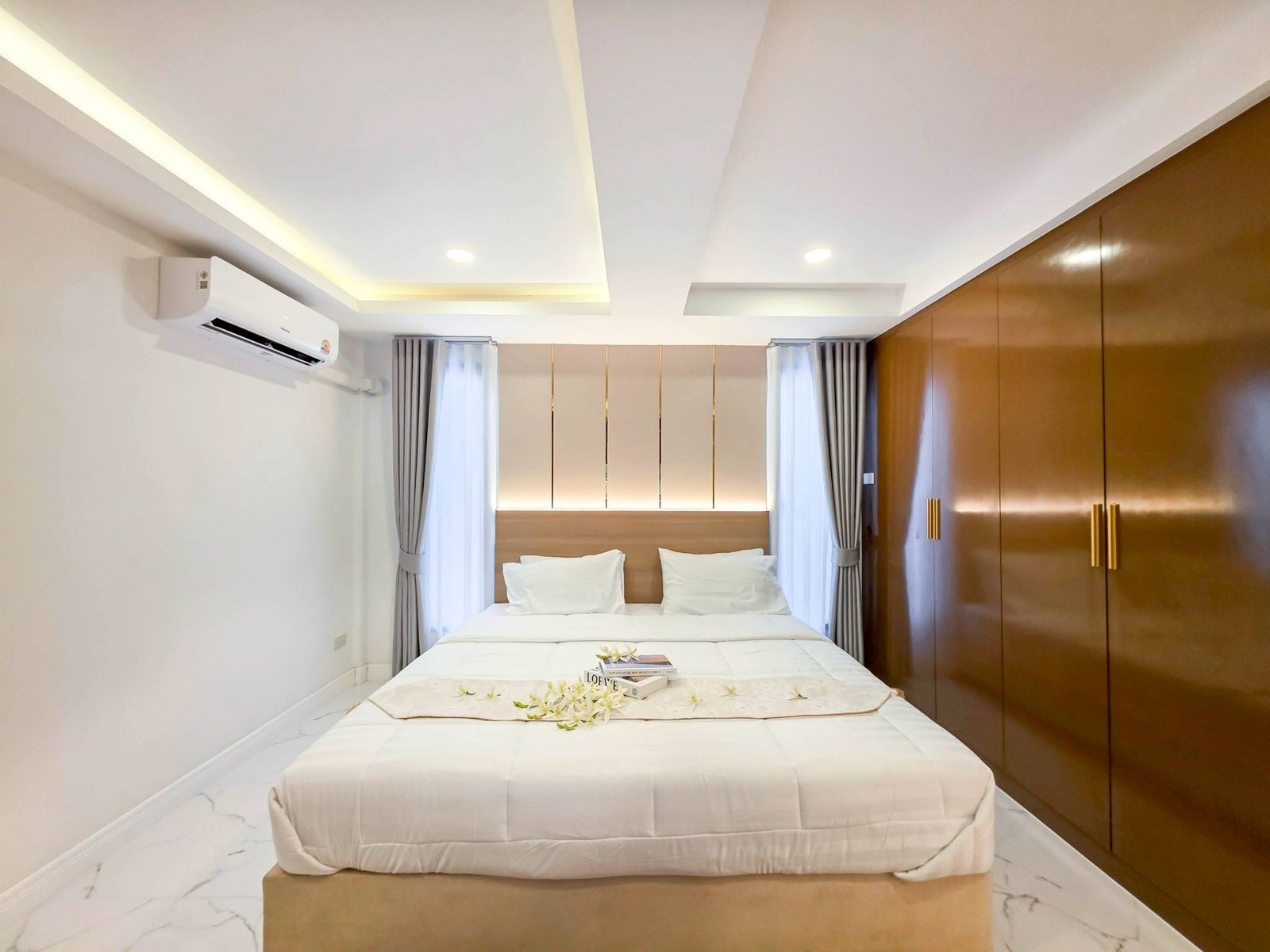 Pool_Villa_South_Pattaya_Sale_Dlh498_7 Hotel room with a large bed, white linens, and a flower-decorated runner; wooden wardrobe on the right and gray curtains.