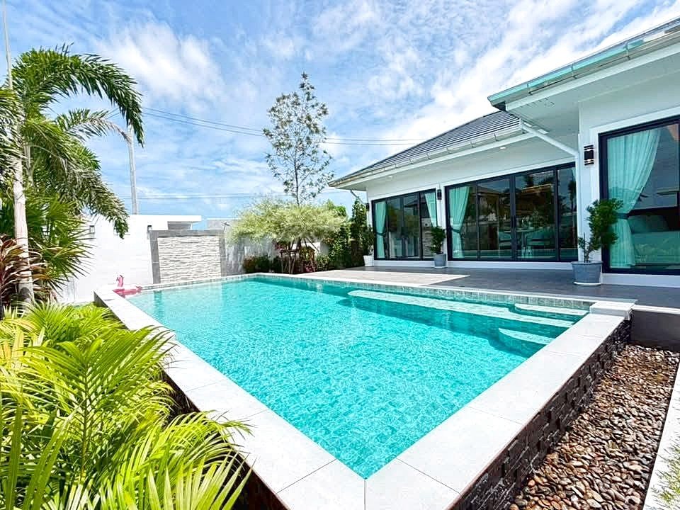 Modern backyard with a rectangular blue pool beside a glass-walled house and tropical plants.