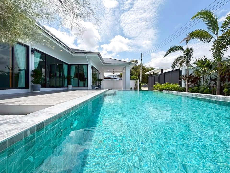 Rectangular turquoise swimming pool beside a modern white house with glass sliding doors and palm trees