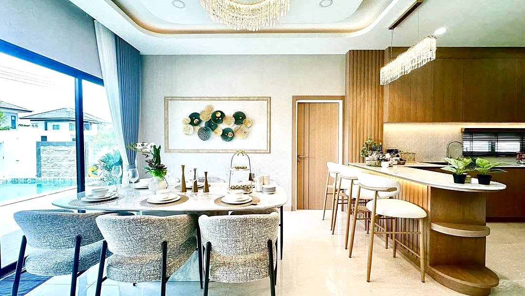 Bright open-plan dining area with a marble table set for six, gray upholstered chairs, and a nearby island with white stools under a crystal chandelier, by large windows and modern wall art.