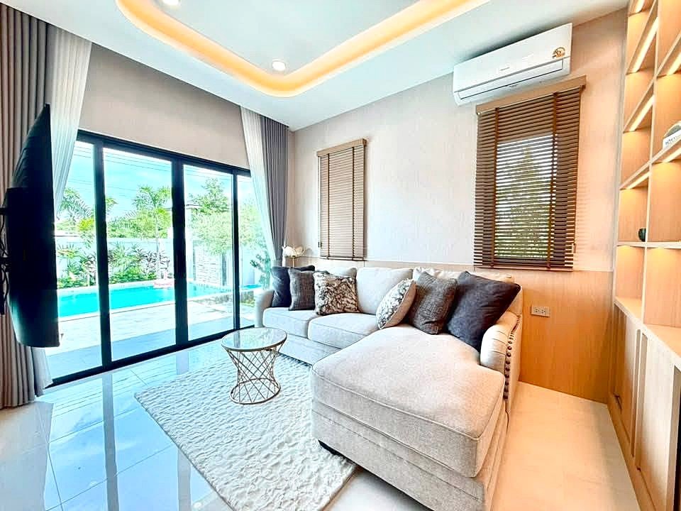 Bright modern living room with a beige sectional sofa, throw pillows, and a glass coffee table, sliding glass doors to a pool area outside.