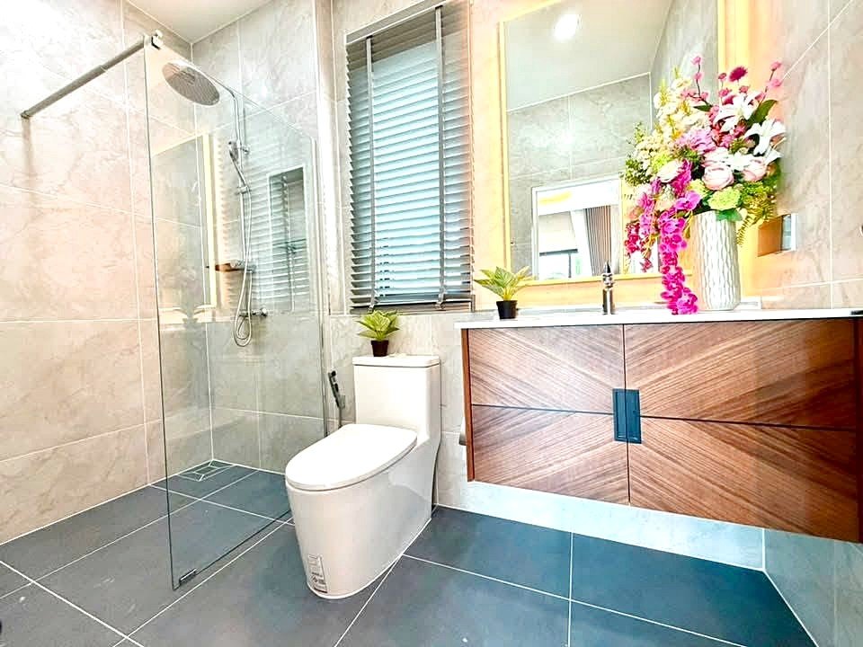 Modern bathroom with glass-enclosed shower, toilet, and wooden vanity topped by a large mirror and colorful flower arrangement, window with blinds to the side.