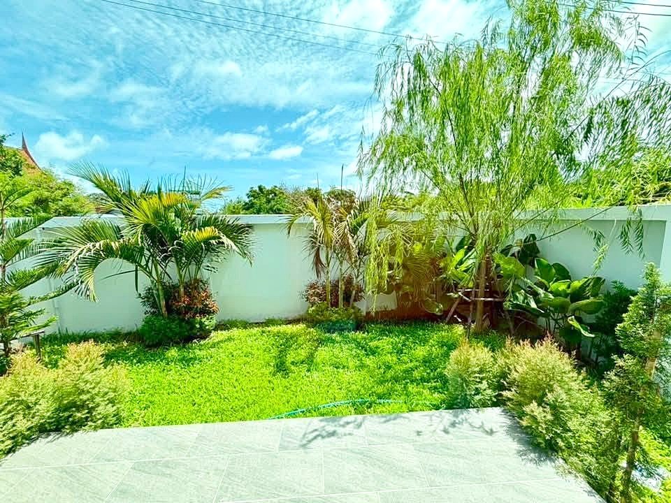 Sunny backyard garden with green lawn, palm trees, and tropical plants along a white fence under a bright blue sky.