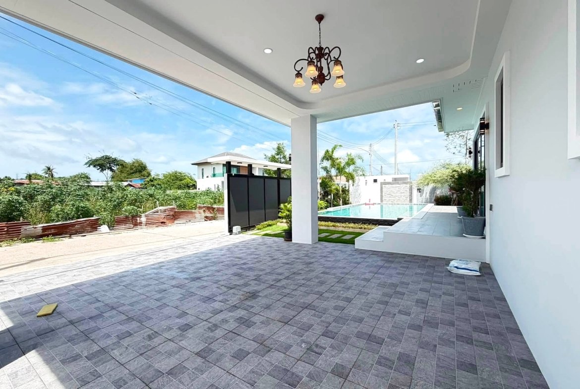 Outdoor covered patio with a bronze chandelier, gray tiled floor, white walls, and a pool beyond under a blue sky.