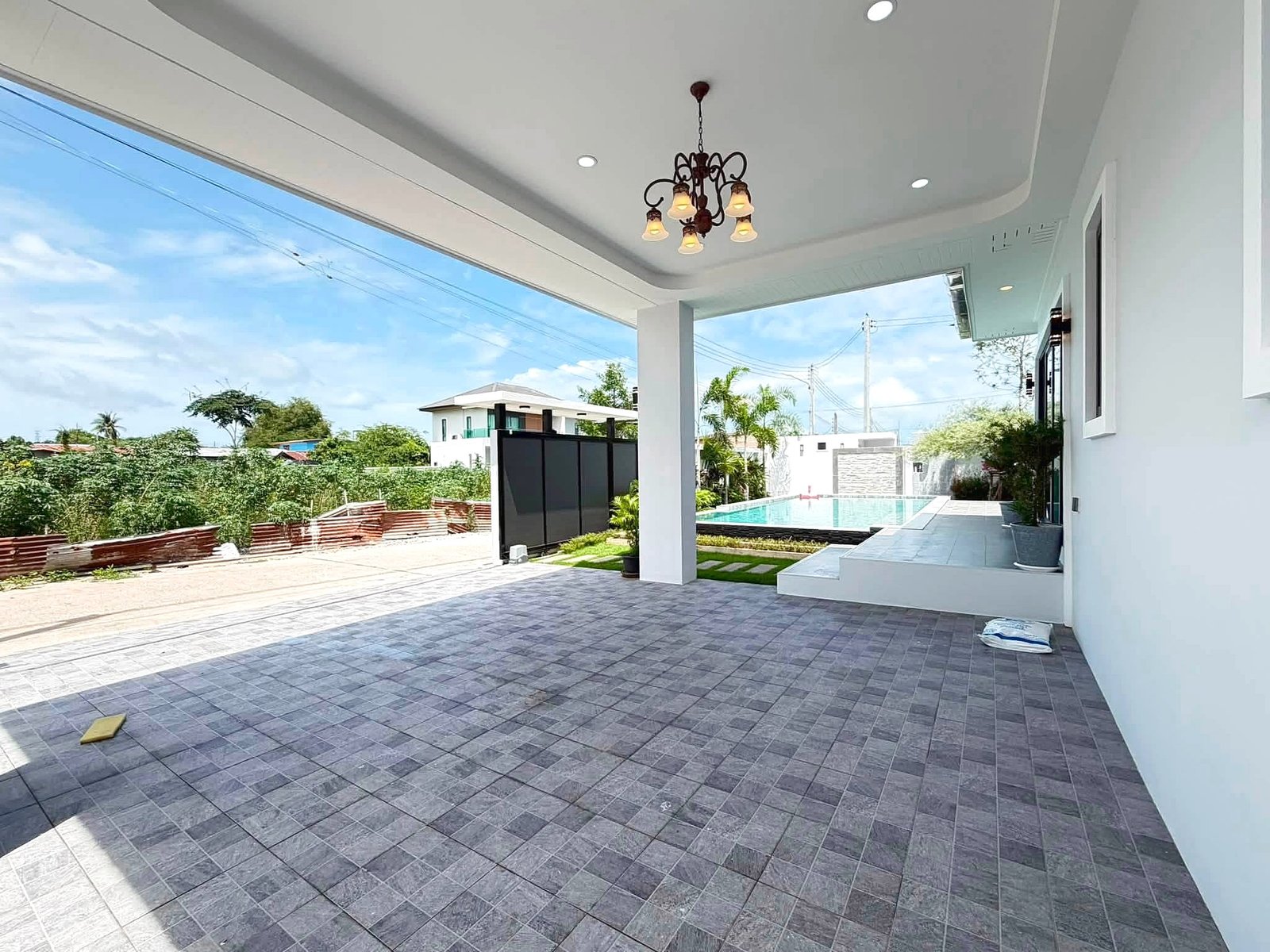 Poolvilla_Khaomakok_Pattaya_Dlh583_28 Outdoor covered patio with a bronze chandelier, gray tiled floor, white walls, and a pool beyond under a blue sky.