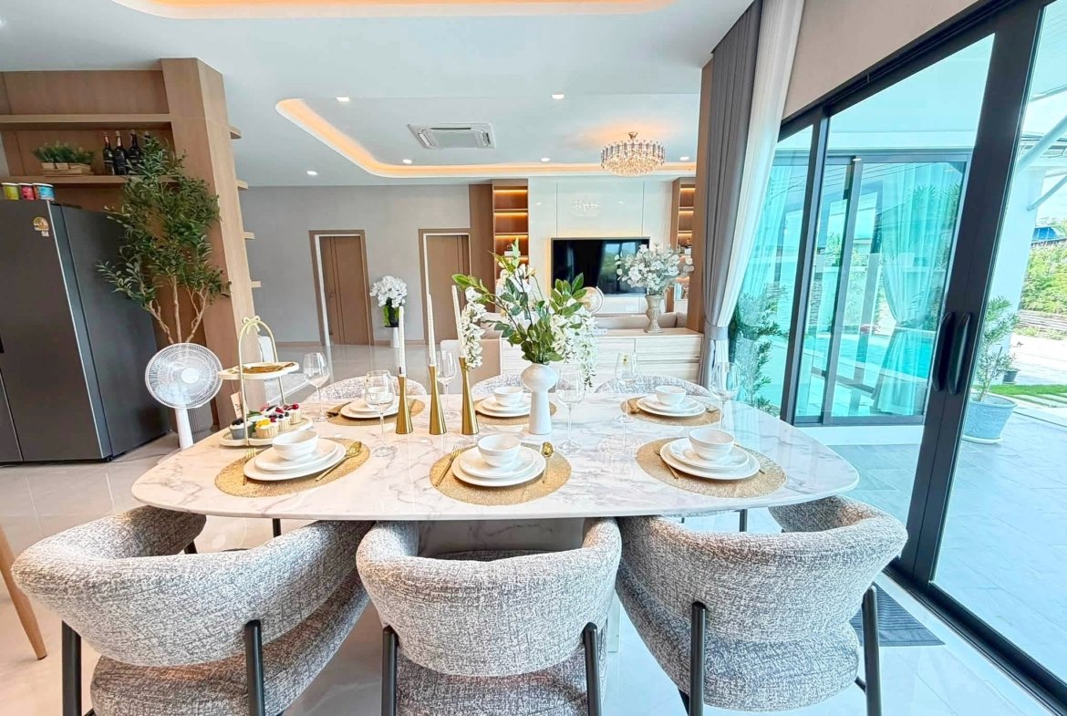 Round marble dining table set for eight with white dishes, glasses, and gold candle holders in a bright, modern room with gray upholstered chairs and a chandelier nearby.