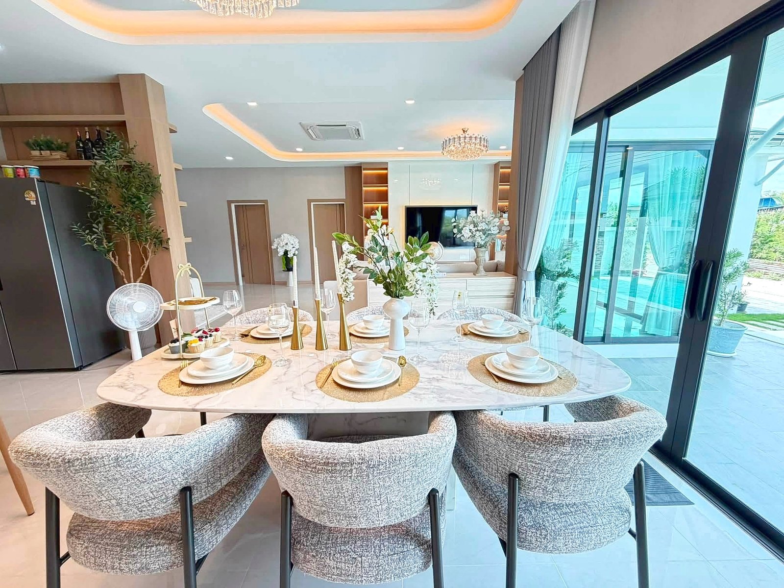 Poolvilla_Khaomakok_Pattaya_Dlh583_4 Round marble dining table set for eight with white dishes, glasses, and gold candle holders in a bright, modern room with gray upholstered chairs and a chandelier nearby.