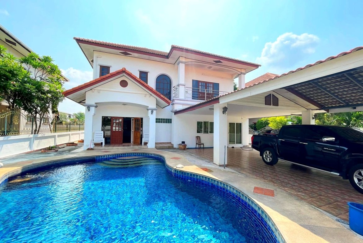 Two-story white house with red-tiled roof beside a curved blue swimming pool; a black SUV parked under a carport on the right, sunny day.