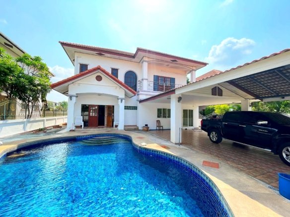 Two story white house with red tiled roof beside a curved blue swimming pool a black SUV parked under a carport on the right sunny day