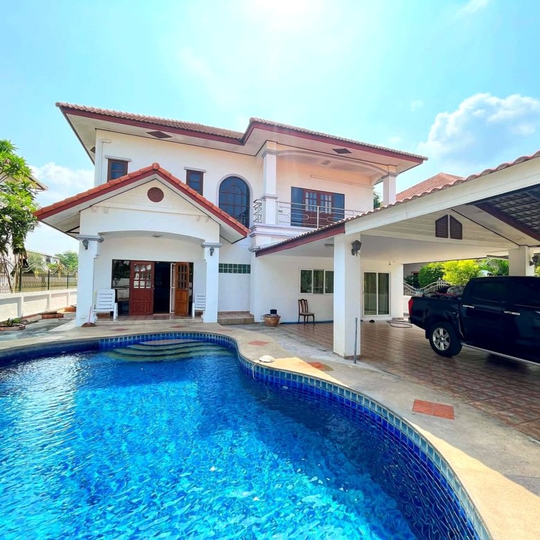 Two story white house with red tiled roof beside a curved blue swimming pool a black SUV parked under a carport on the right sunny day