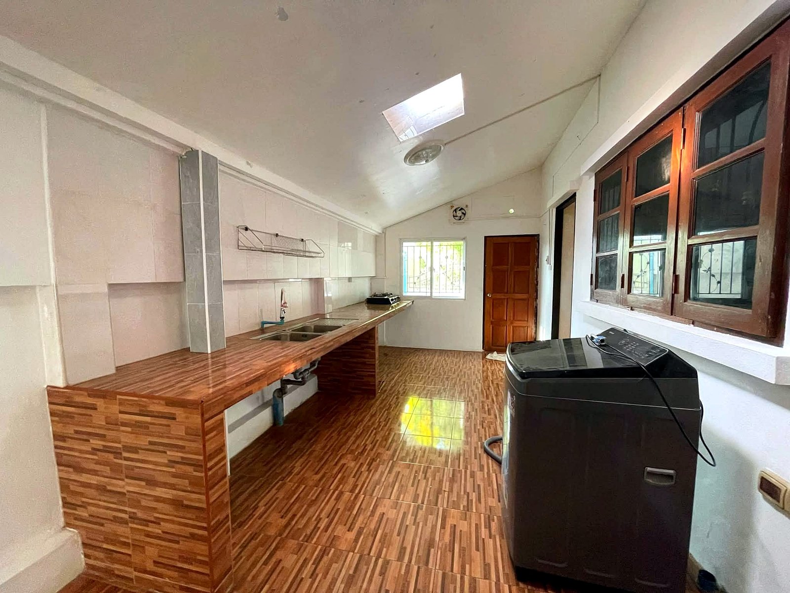 Poolvilla_Pattaya_Rent_Rdl352_10 Bright kitchen with a long wooden island and dual sinks, white cabinets, and a skylight; a dark washing machine is in the foreground near the windowed wall on the right.