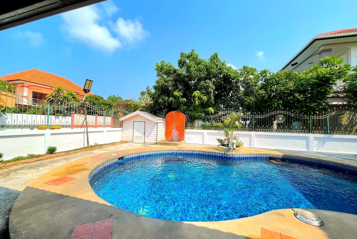 Outdoor swimming pool with blue water, turquoise tiles, and surrounding concrete deck amid a white fence and tropical trees under a clear blue sky