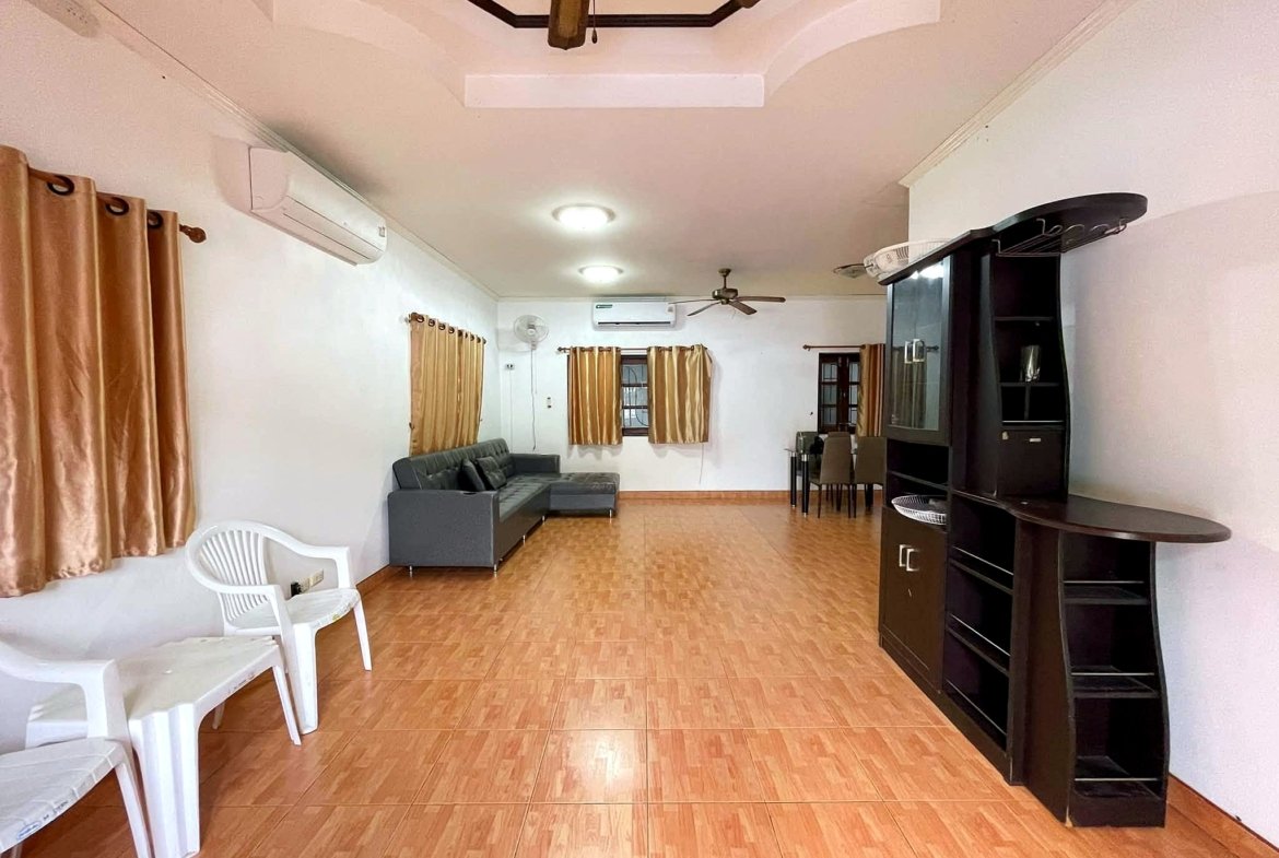 Living room with tan curtains, brown tiled floor, gray sectional sofa, white plastic chairs, and a tall dark cabinet along the right wall.