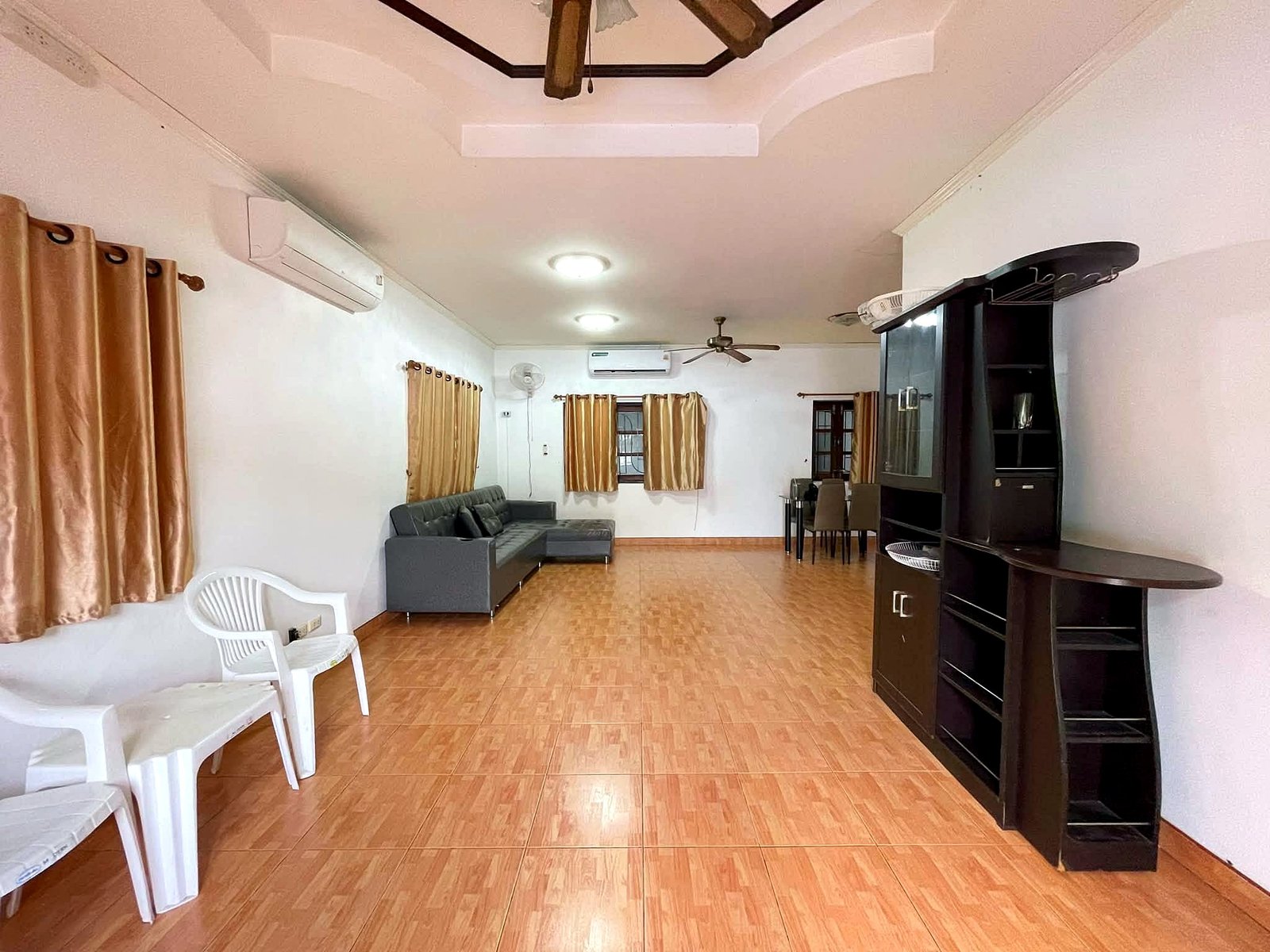 Poolvilla_Pattaya_Rent_Rdl352_3 Living room with tan curtains, brown tiled floor, gray sectional sofa, white plastic chairs, and a tall dark cabinet along the right wall.