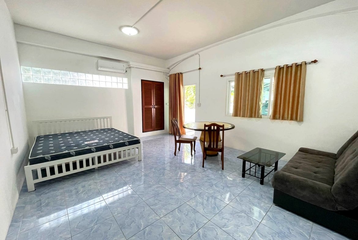 Bright living area with a bed on the left, a wooden wardrobe, round glass dining table and beige curtains by windows.