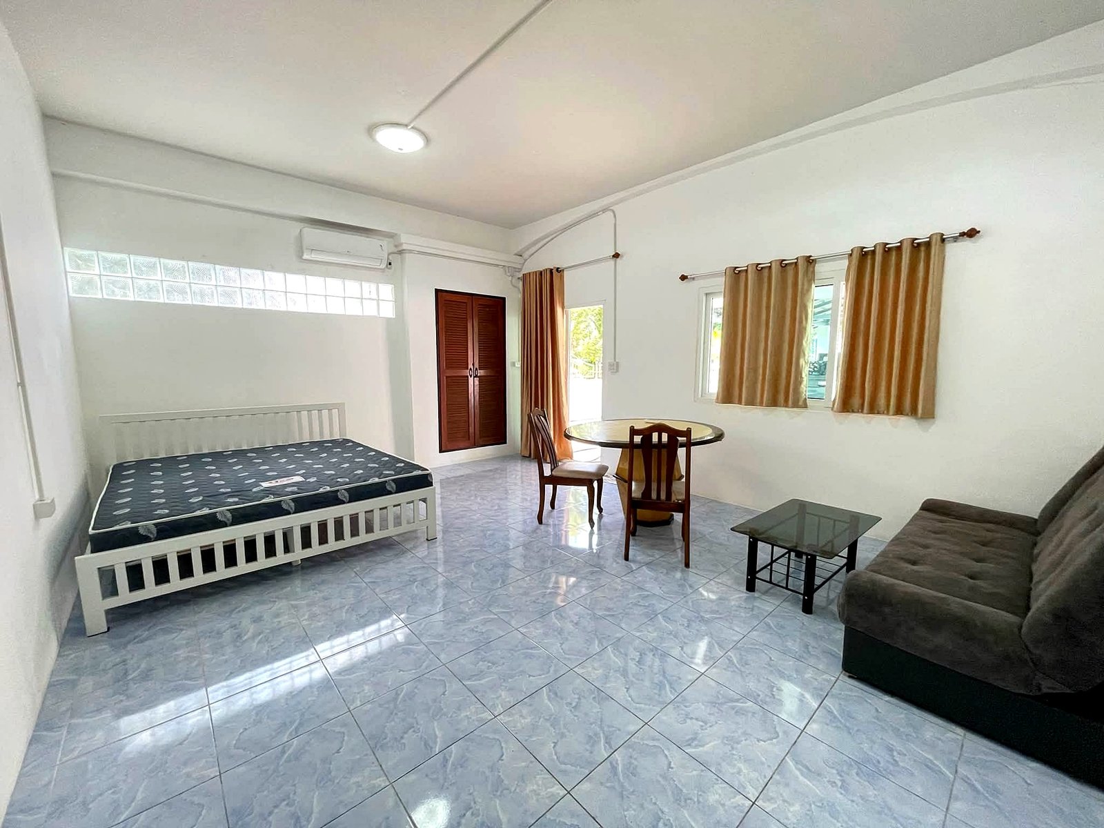 Poolvilla_Pattaya_Rent_Rdl352_5 Bright living area with a bed on the left, a wooden wardrobe, round glass dining table and beige curtains by windows.