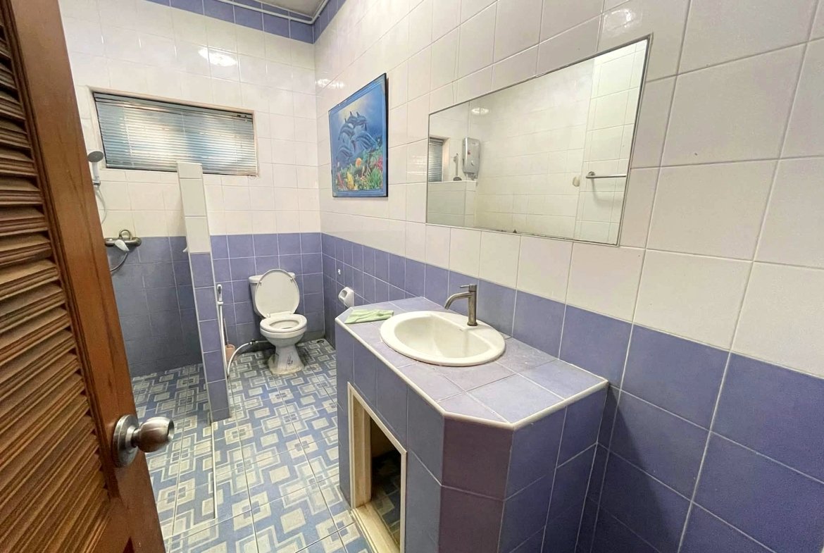Blue-tiled bathroom with a white sink, toilet, and a large rectangular mirror; ocean-themed painting on the wall and a window with blinds.