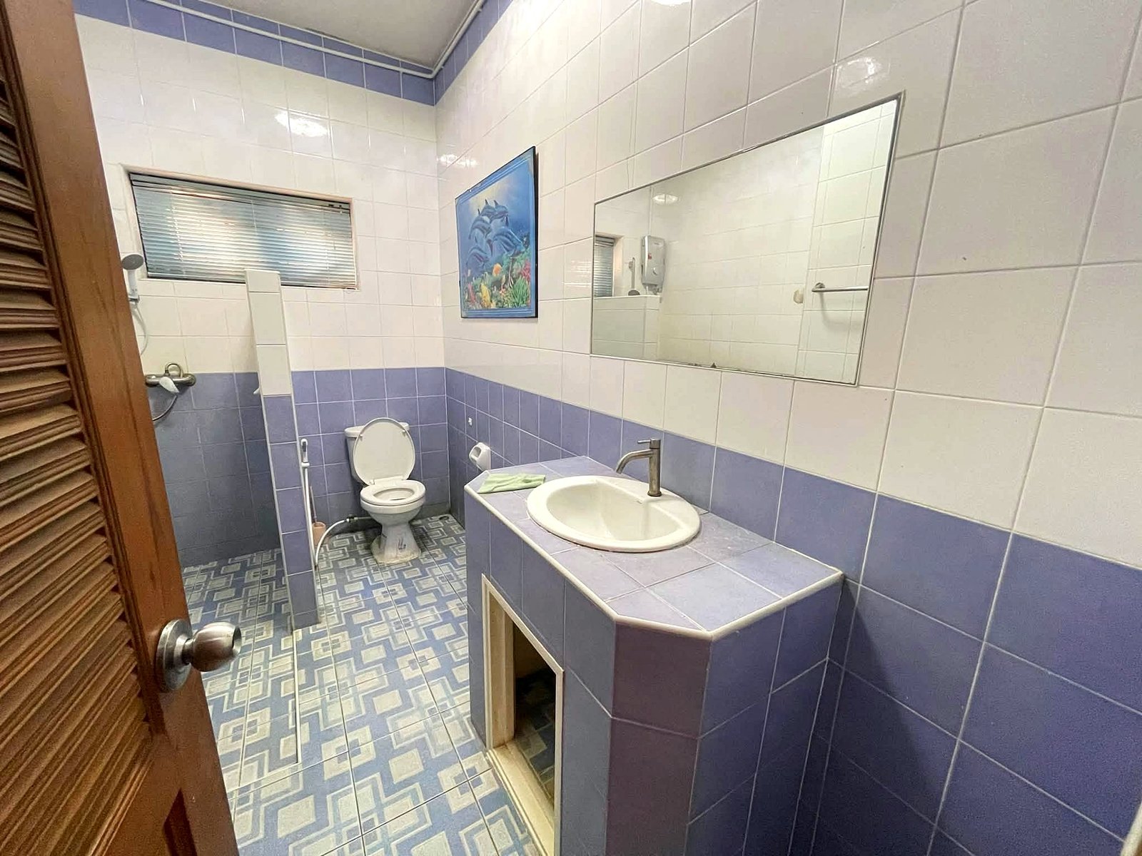 Poolvilla_Pattaya_Rent_Rdl352_6 Blue-tiled bathroom with a white sink, toilet, and a large rectangular mirror; ocean-themed painting on the wall and a window with blinds.