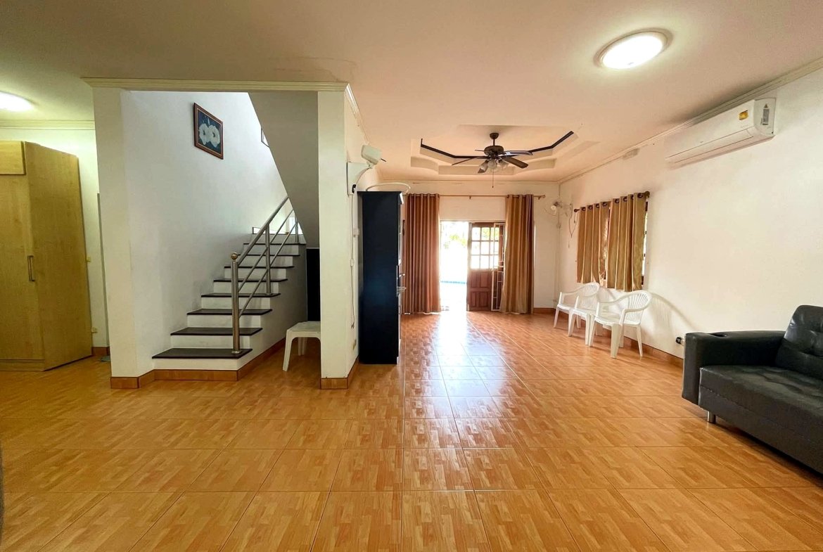 Spacious living room with staircase on the left, ceiling fan, and brown tiled floor; white chairs along the right wall near windows with curtains.