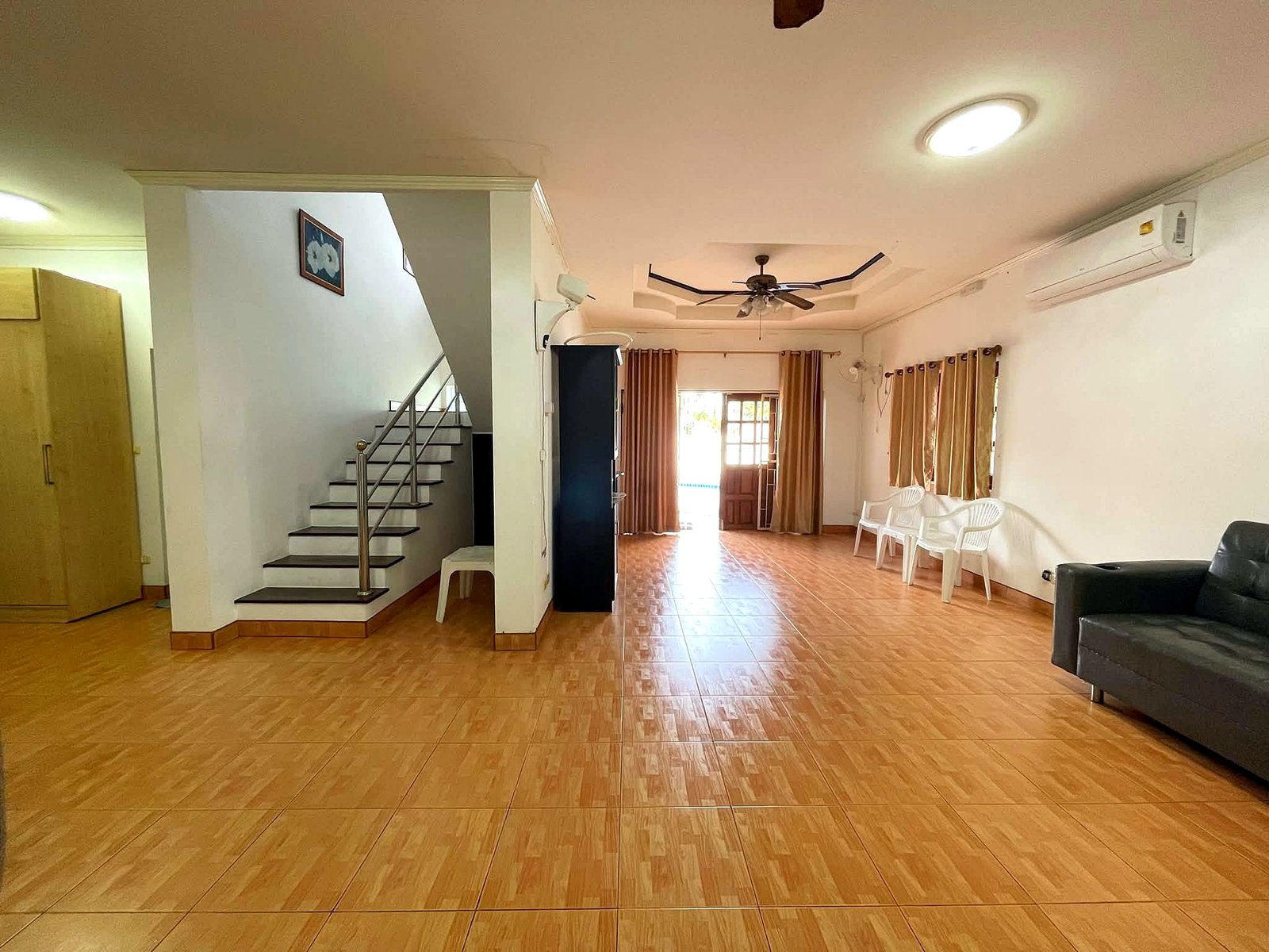 Poolvilla_Pattaya_Rent_Rdl352_7 Spacious living room with staircase on the left, ceiling fan, and brown tiled floor; white chairs along the right wall near windows with curtains.