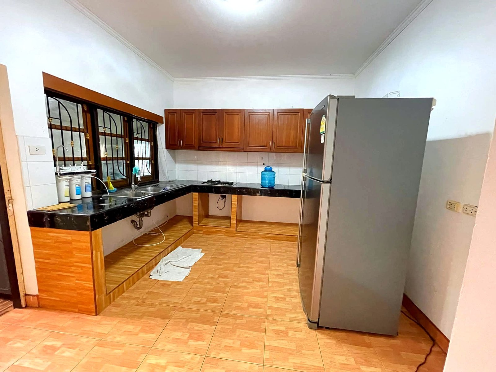 Poolvilla_Pattaya_Rent_Rdl352_9 Kitchen with brown wooden cabinets, black countertops, a stainless steel refrigerator, sink, and gas stove; tiled floor and barred window on the left.