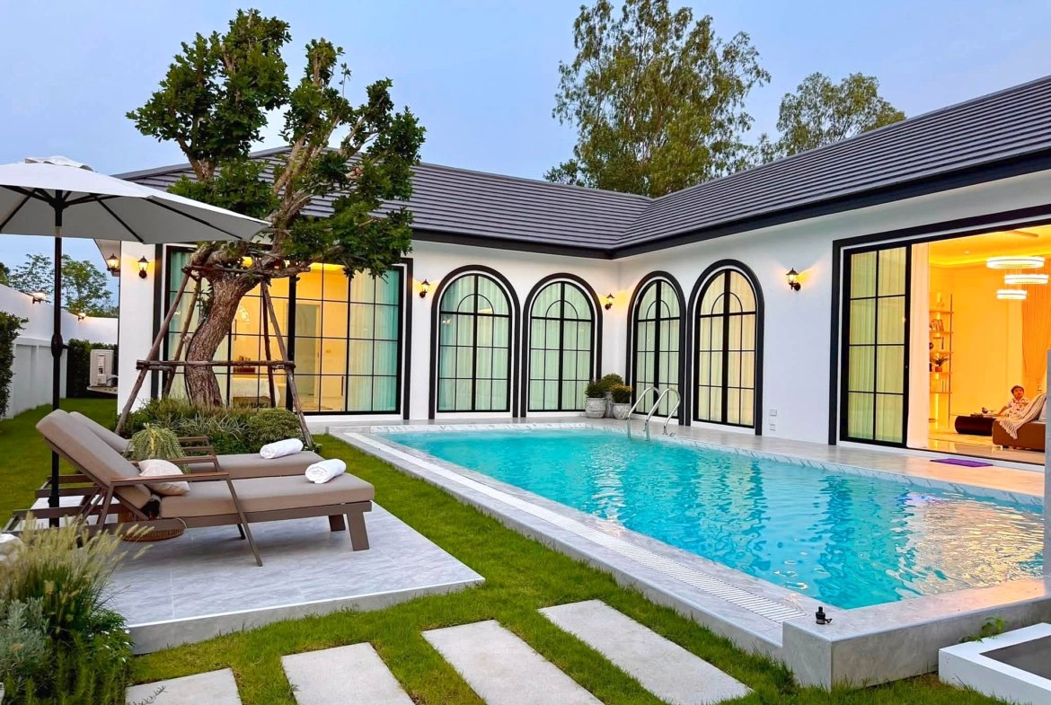 Modern backyard with a rectangular pool, stepping-stone path, brown lounge chairs under a white umbrella, and a tree, adjacent to a white house with large arched windows. Interior glow visible through doors.