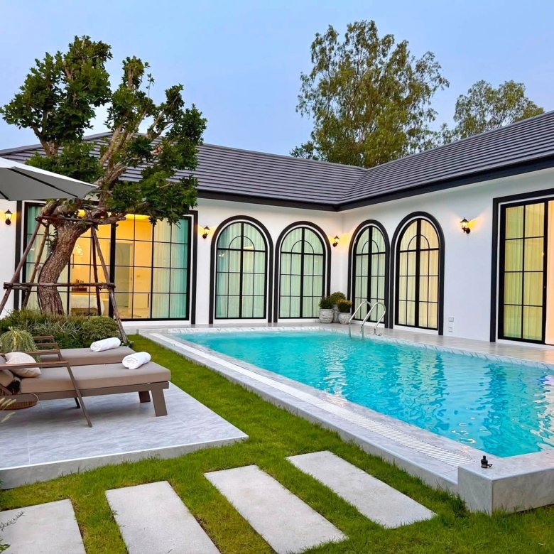 Modern backyard with a rectangular pool stepping stone path brown lounge chairs under a white umbrella and a tree adjacent to a white house with large arched windows Interior glow visible through doors