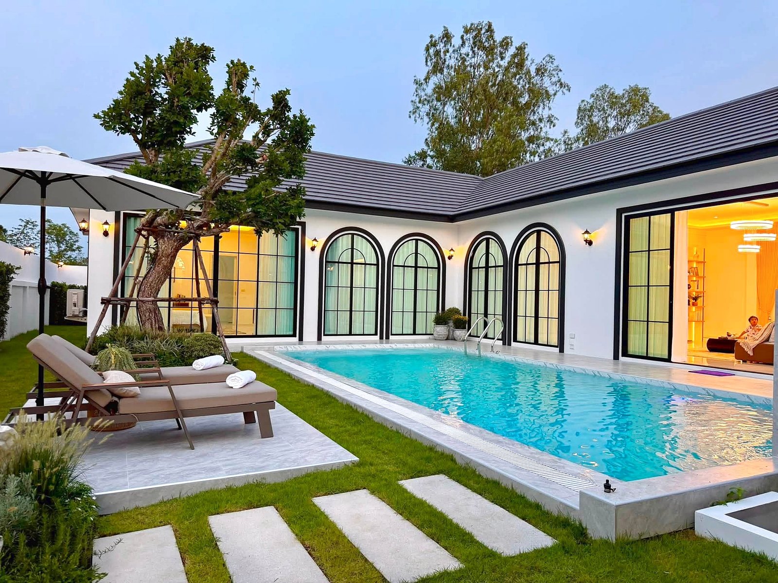 Poolvilla_Soi_Siam_Pattaya_Dlh584 Modern backyard with a rectangular pool, stepping-stone path, brown lounge chairs under a white umbrella, and a tree, adjacent to a white house with large arched windows. Interior glow visible through doors.