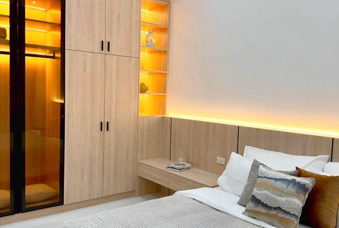 Modern bedroom with light wood wardrobes, glowing open shelves, and a neatly made bed with a gray striped blanket and decorative pillows.
