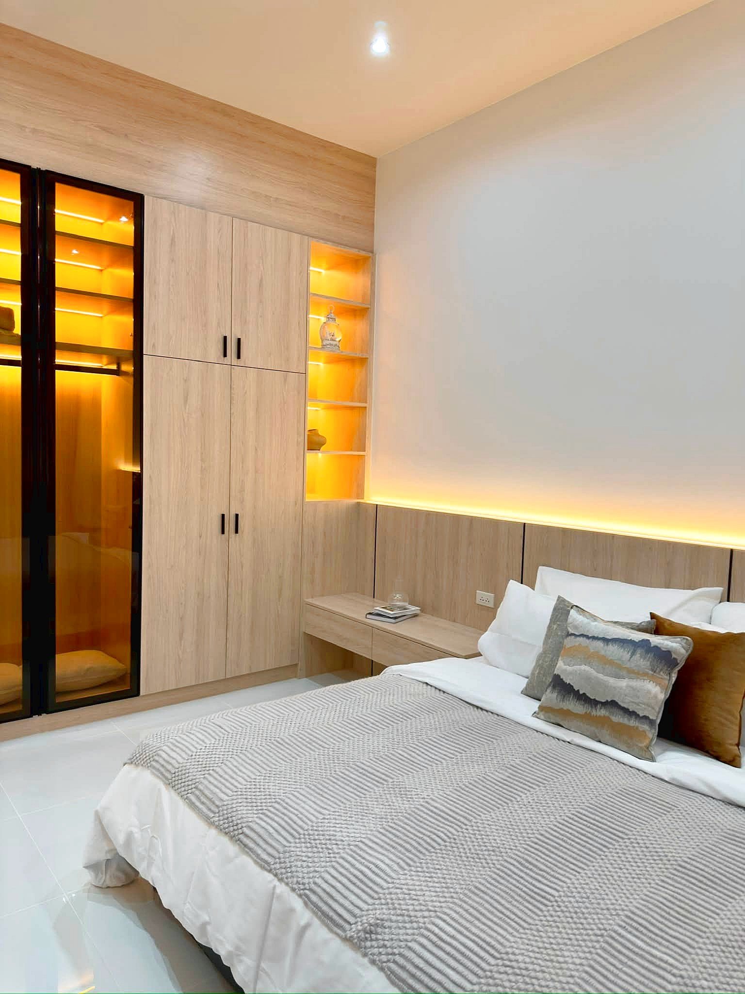 Poolvilla_Soi_Siam_Pattaya_Dlh584_28 Modern bedroom with light wood wardrobes, glowing open shelves, and a neatly made bed with a gray striped blanket and decorative pillows.