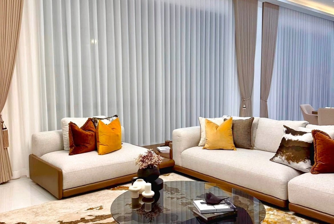 Modern living room with a cream sectional, orange pillows, and a round glass coffee table on a gold-toned rug under soft lighting.