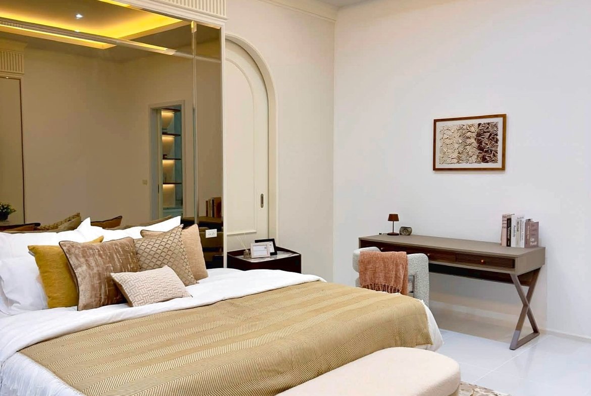 Hotel-style bedroom with beige-gold bed linens and multiple throw pillows, a white bench at the foot, and a patterned rug.