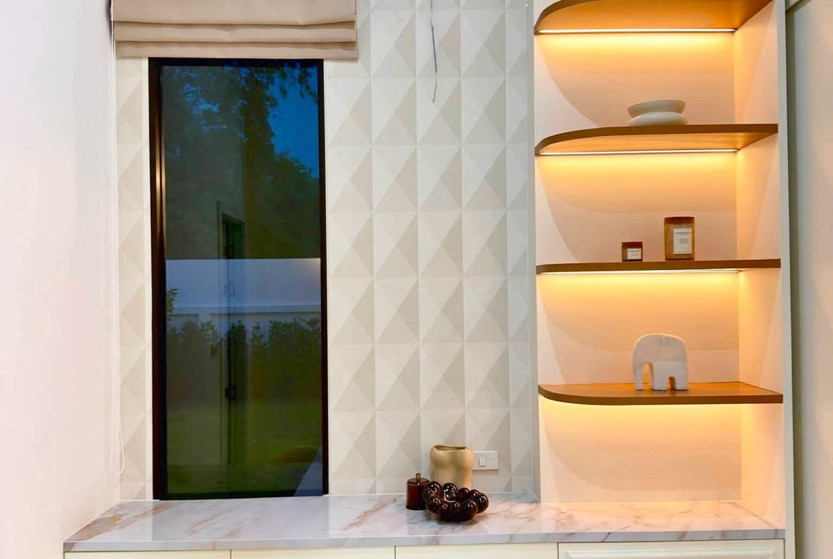 Cream-toned bathroom vanity area with built-in illuminated shelves on the right, beige window shade to the left, and a marble-topped counter with a beige upholstered stool in front.