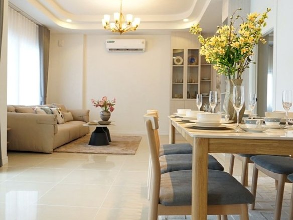 Bright open-plan living and dining room with a wooden table set for six, a vase of yellow flowers, and a beige sofa in the background.