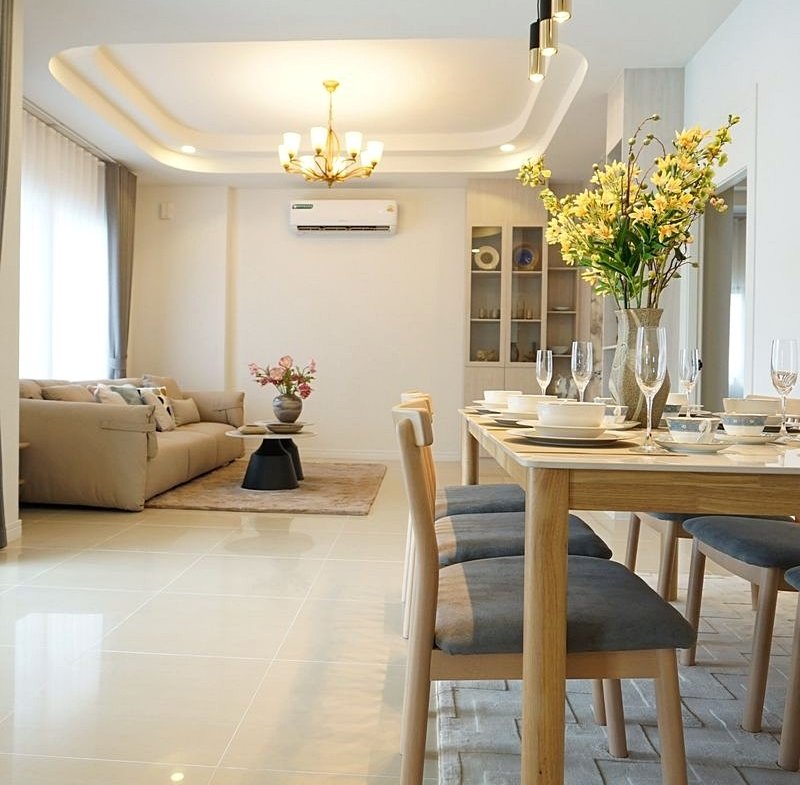 Bright open-plan living and dining room with a wooden table set for six, a vase of yellow flowers, and a beige sofa in the background.
