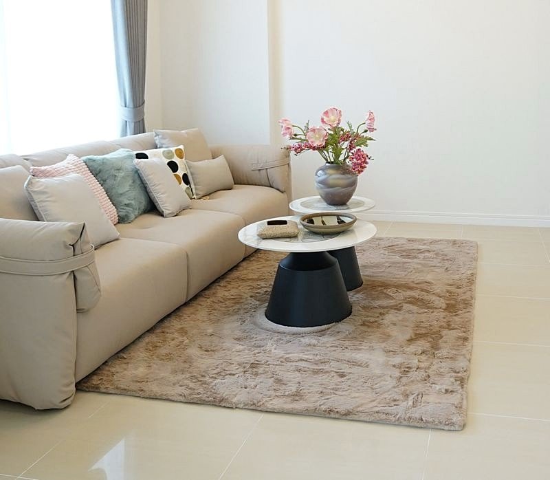 Beige sectional sofa with assorted throw pillows, two round marble coffee tables, and a vase of pink flowers on a beige rug in a bright living room.