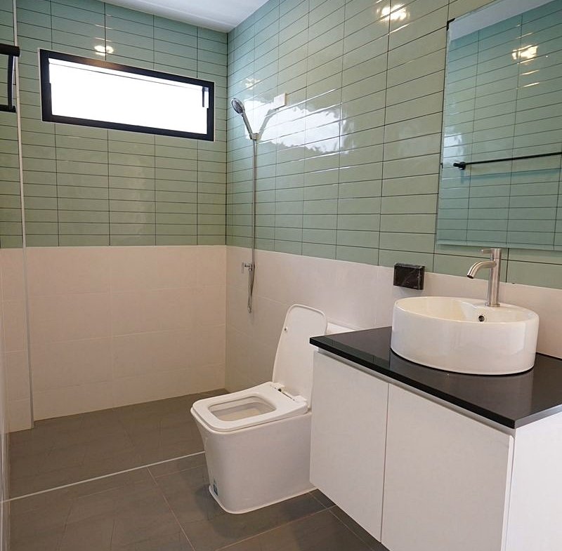 Modern bathroom with pale green wall tiles, white toilet, white vanity with black countertop and round vessel sink, mirror and window above.