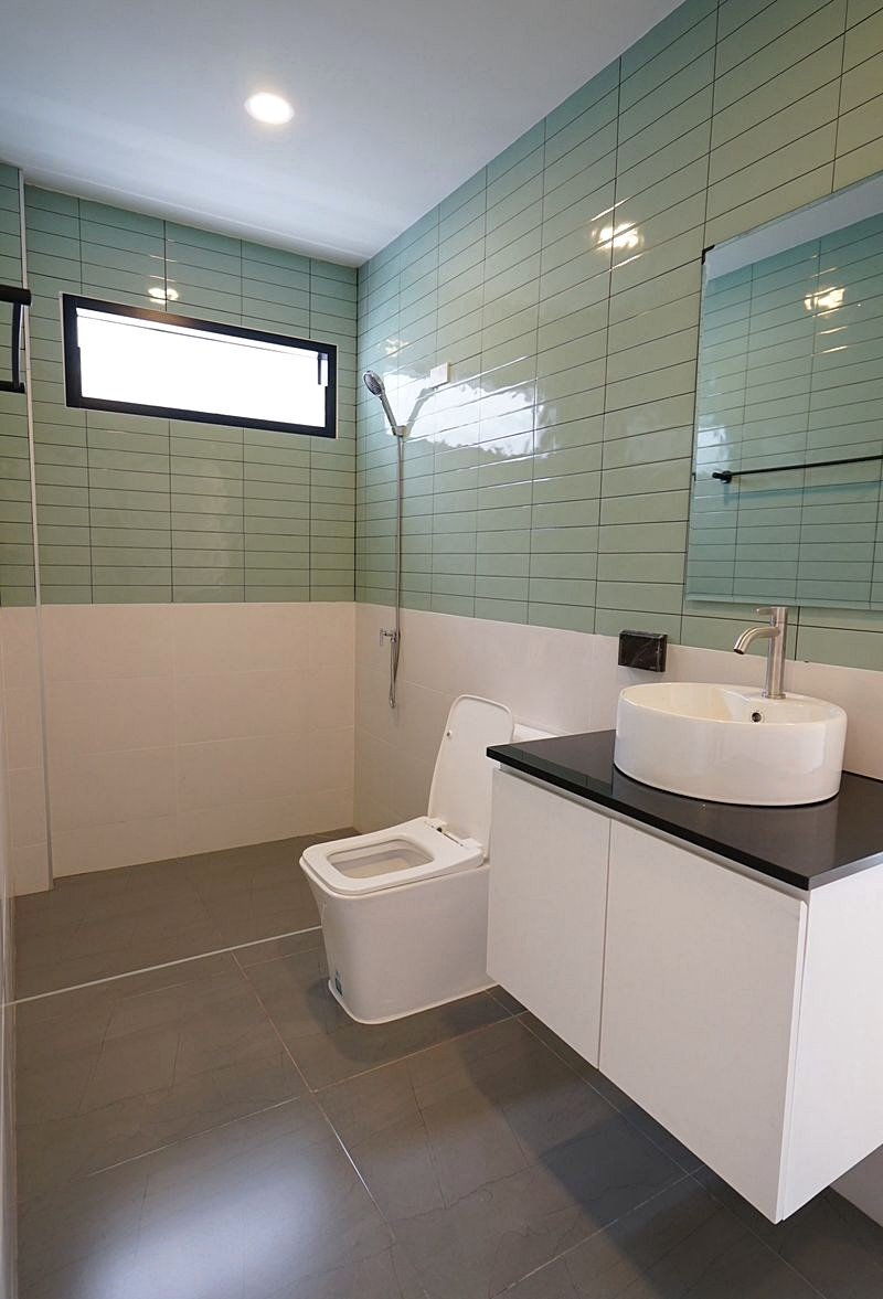 Pornthep_2_Village_Pattaya_Dlh578_8 Modern bathroom with pale green wall tiles, white toilet, white vanity with black countertop and round vessel sink, mirror and window above.